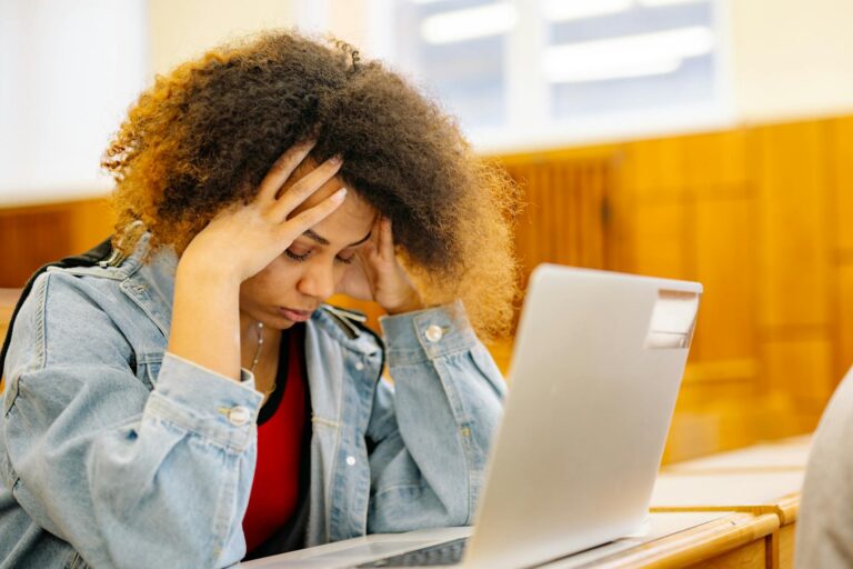 A young woman experiencing a headache while studying with a laptop indoors, looking stressed and overwhelmed.
