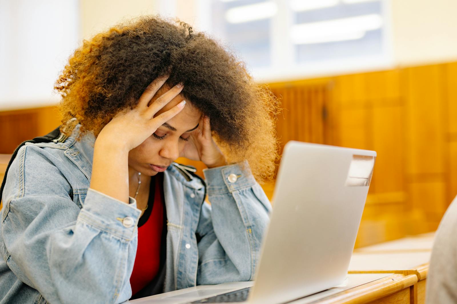 A young woman experiencing a headache while studying with a laptop indoors, looking stressed and overwhelmed.